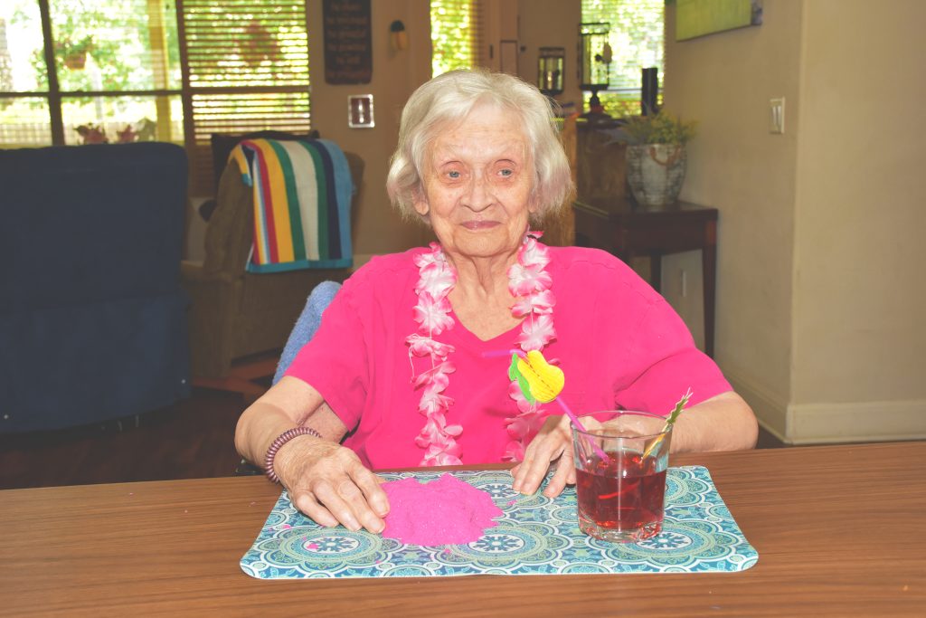 An old woman in a pink shirt is playing with clay on the table with her juice on the side.
